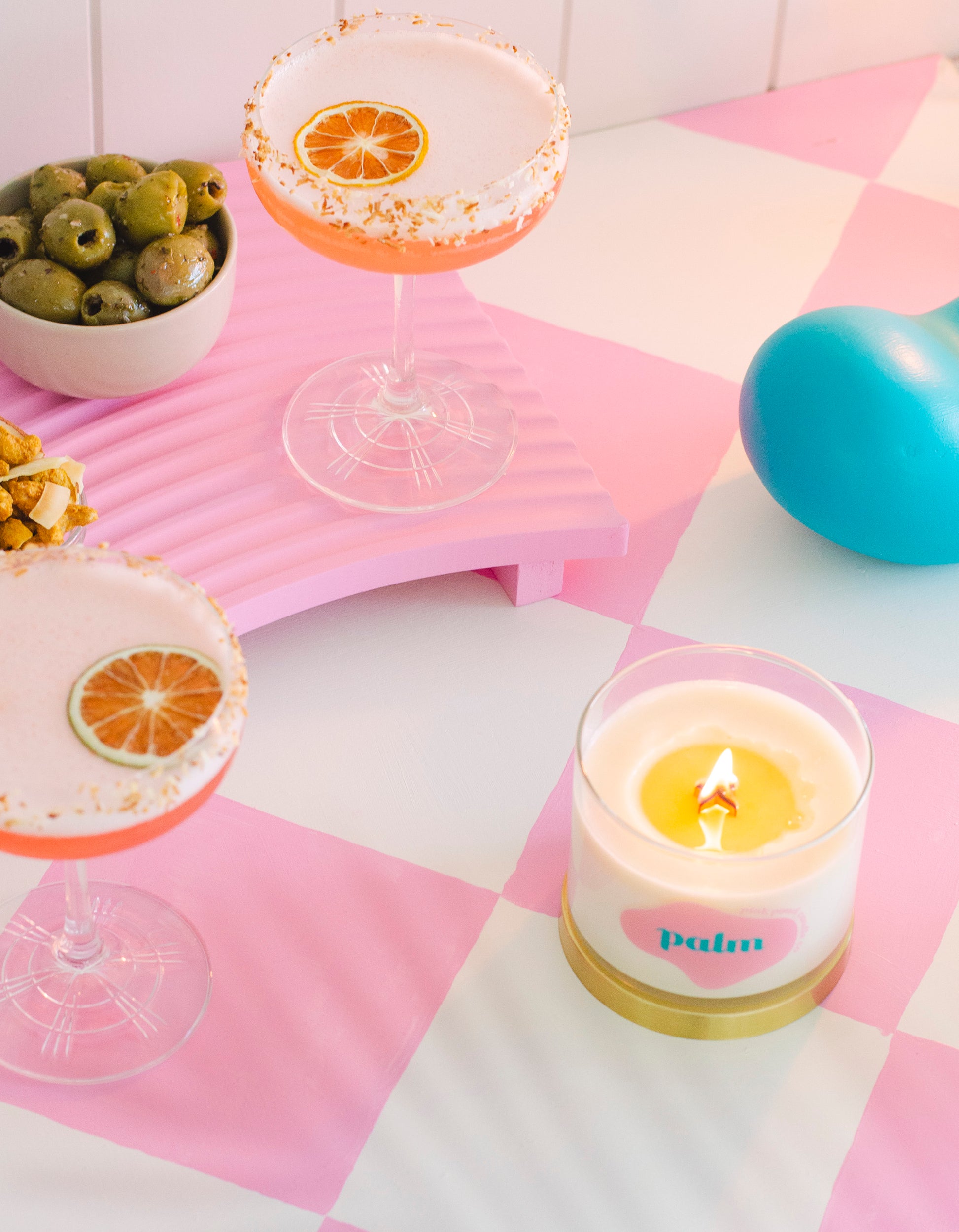 A lit coconut palm candle with cocktails and olives on a pink checker background